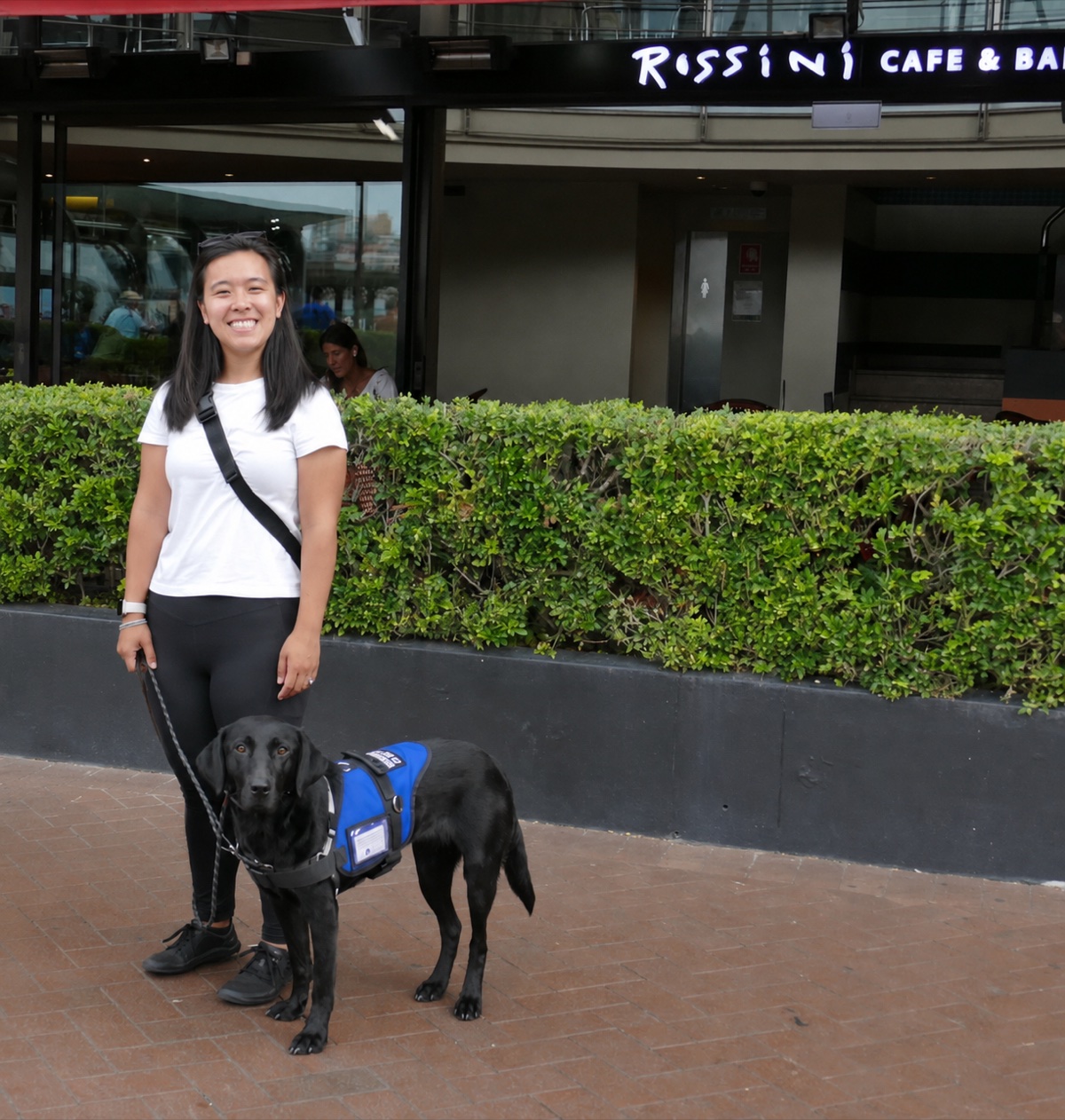 Diona outside a cafe with Murphy, her black Labrador PTSD assistance dog, settled in her blue working vest
